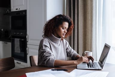 Jonge vrouw werkt aan de keukentafel op een laptop, met papieren en een mok voor zich