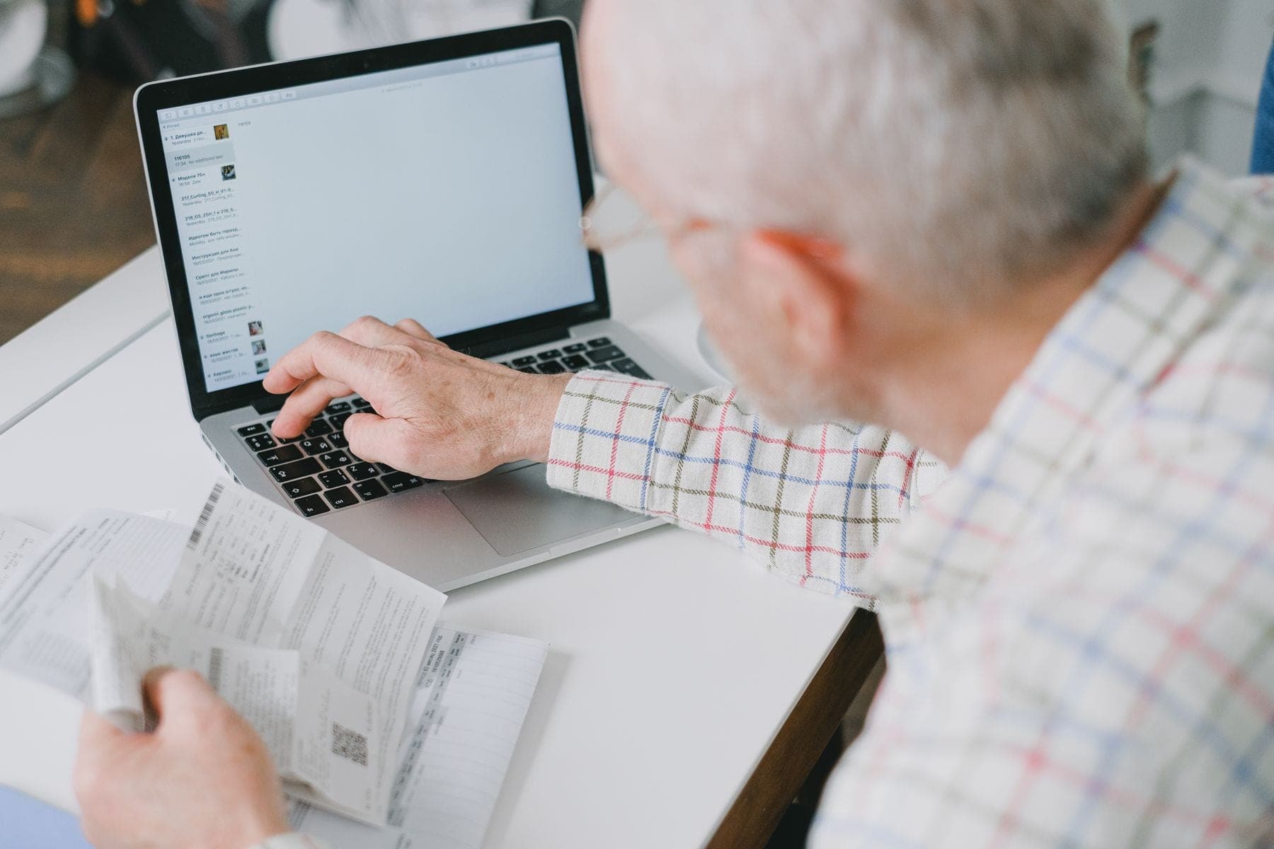 Oudere man aan een bureau bekijkt zijn bankzaken op een laptop, met papieren in de hand
