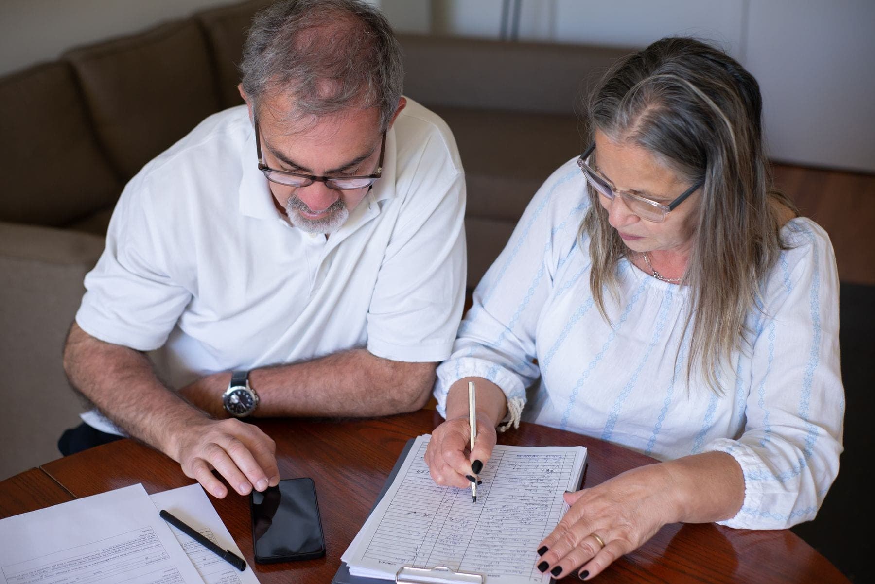 Oudere man en vrouw aan een houten tafel; de vrouw vult met een pen een formulier in, de man kijkt mee