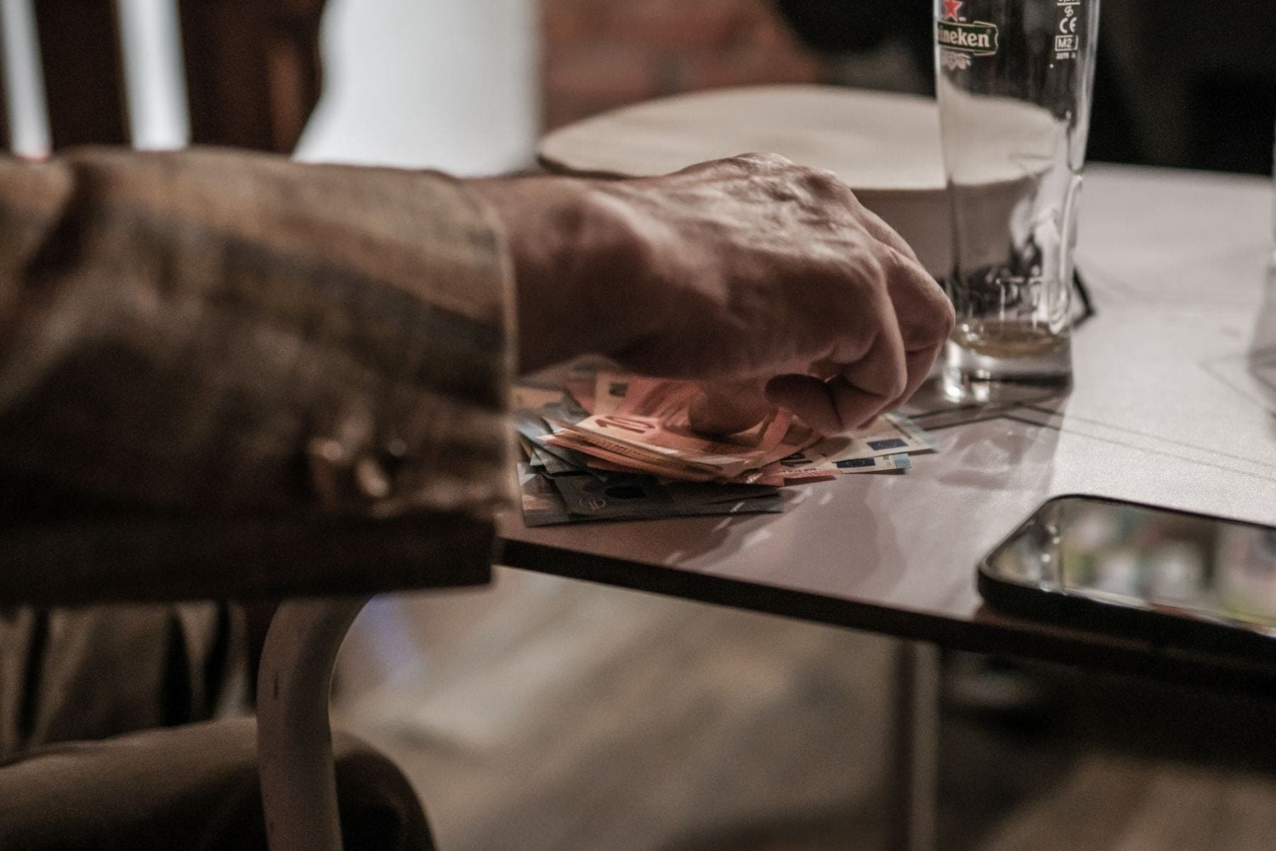 Een hand telt eurobiljetten aan een tafel, naast een telefoon en een leeg glas