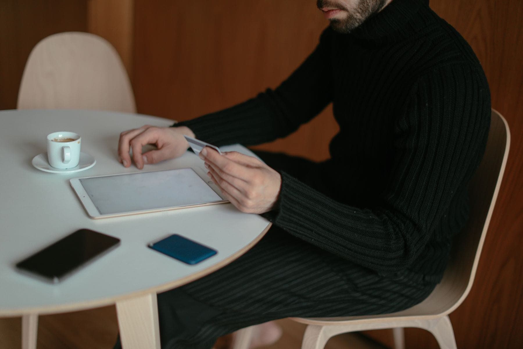 Man aan tafel thuis houdt een bankpas vast en bestelt iets op een tablet, met een kop koffie naast zich