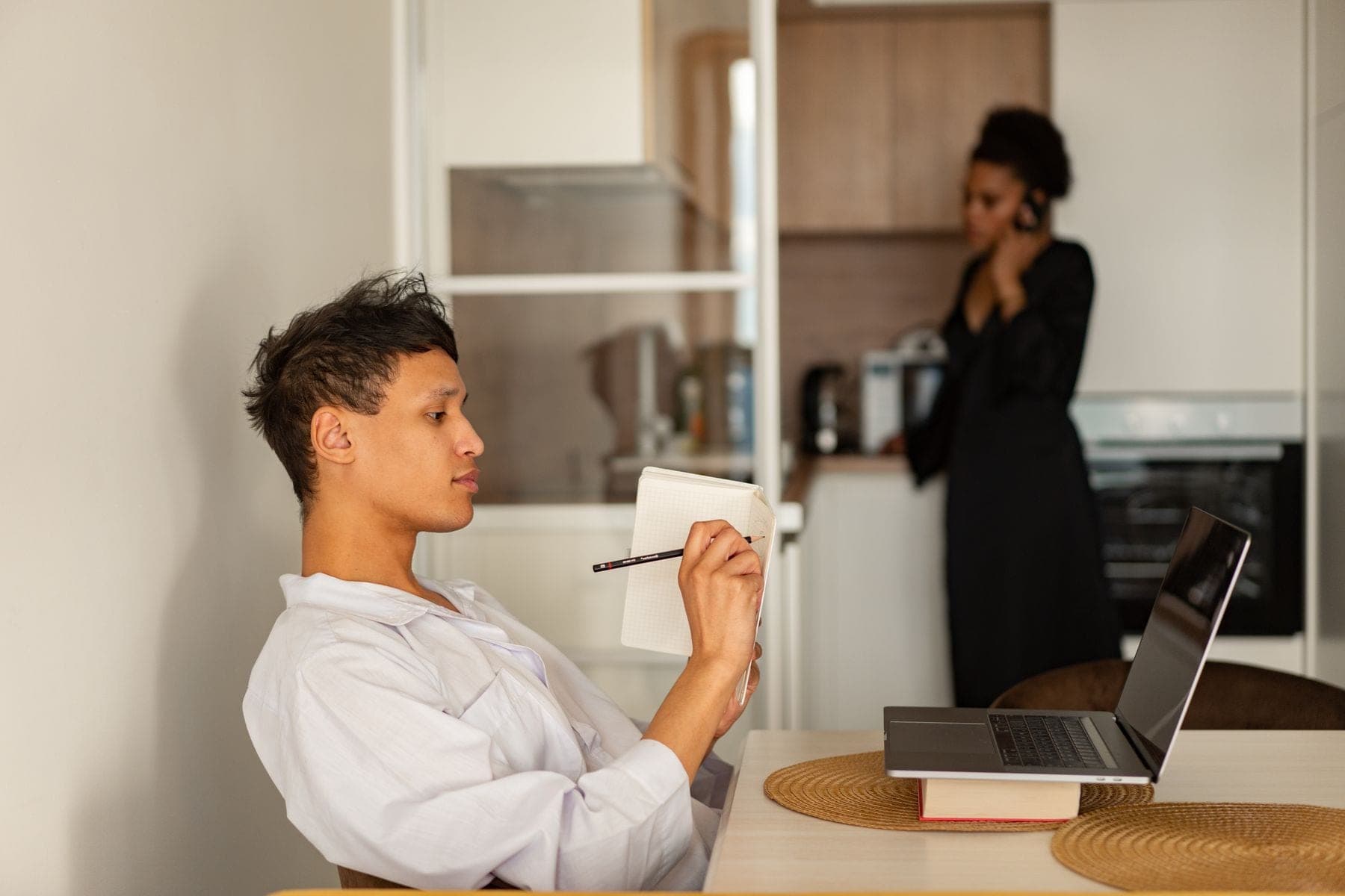 Man schrijft thuis aantekeningen naast een laptop, op de achtergrond belt een vrouw