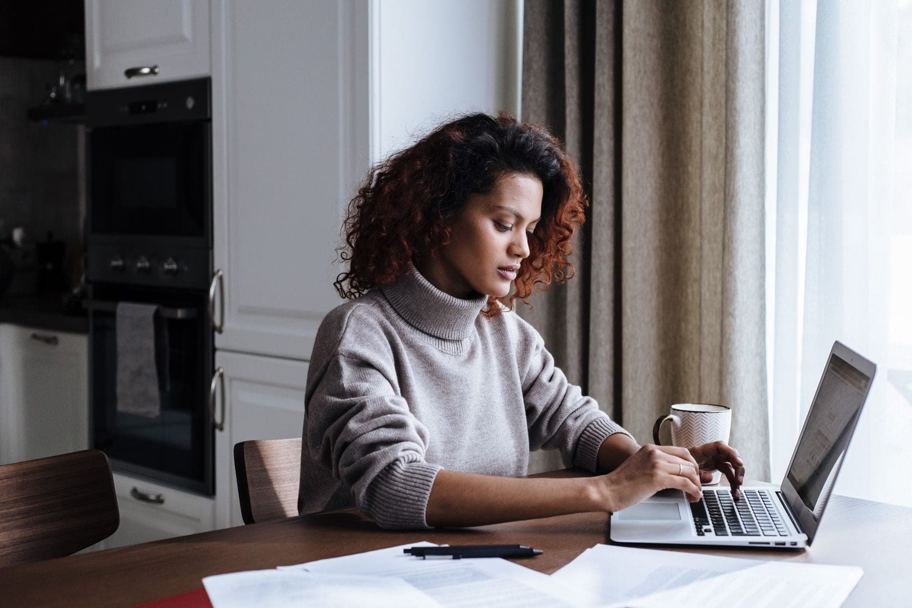 Jonge vrouw werkt aan de keukentafel op een laptop, met papieren en een mok voor zich