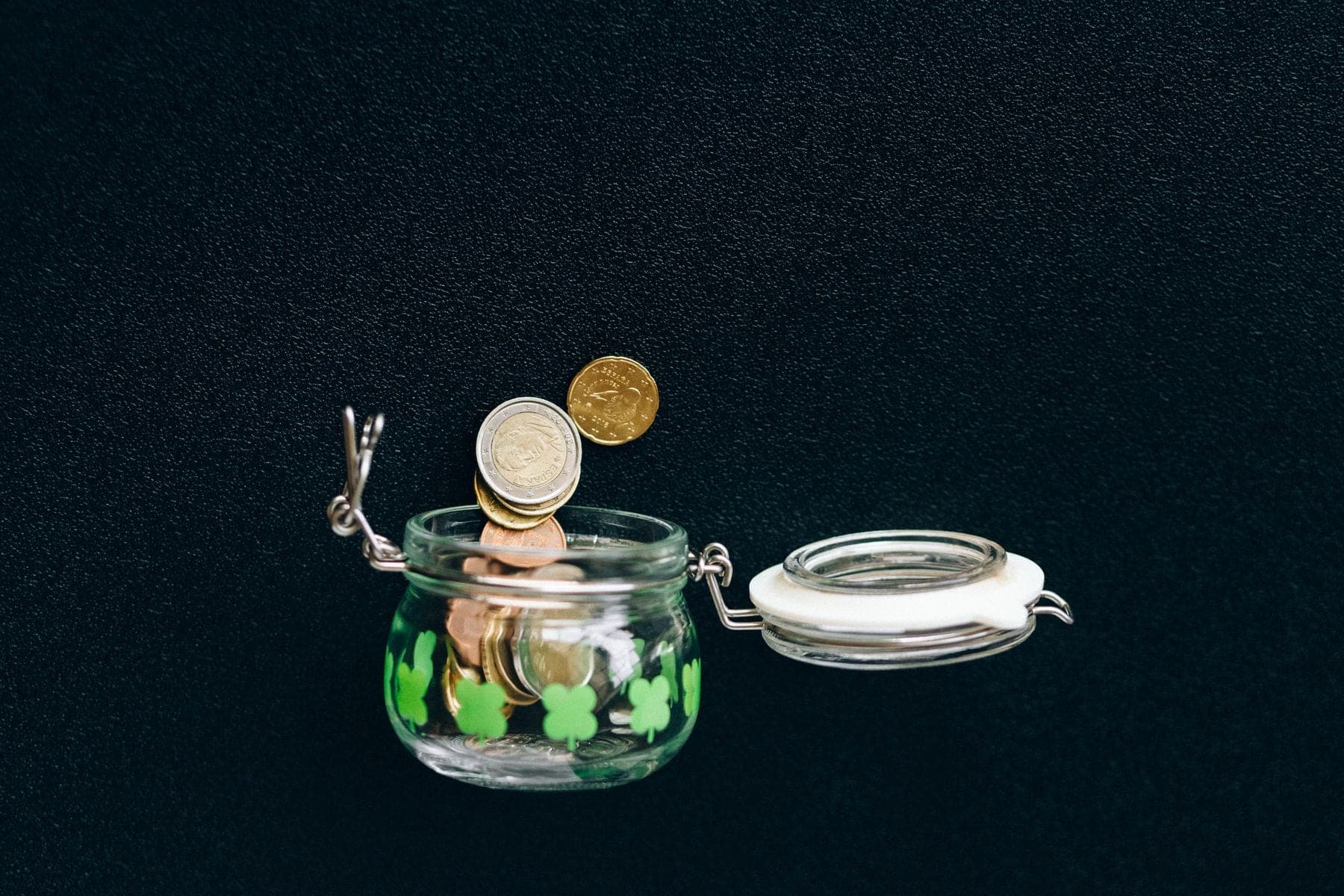 Euromunten vallen in een open glazen weckpot tegen een donkere achtergrond