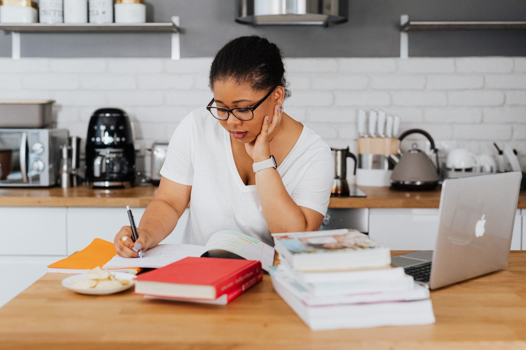 Jonge vrouw schrijft aan een keukentafel met boeken en een open laptop