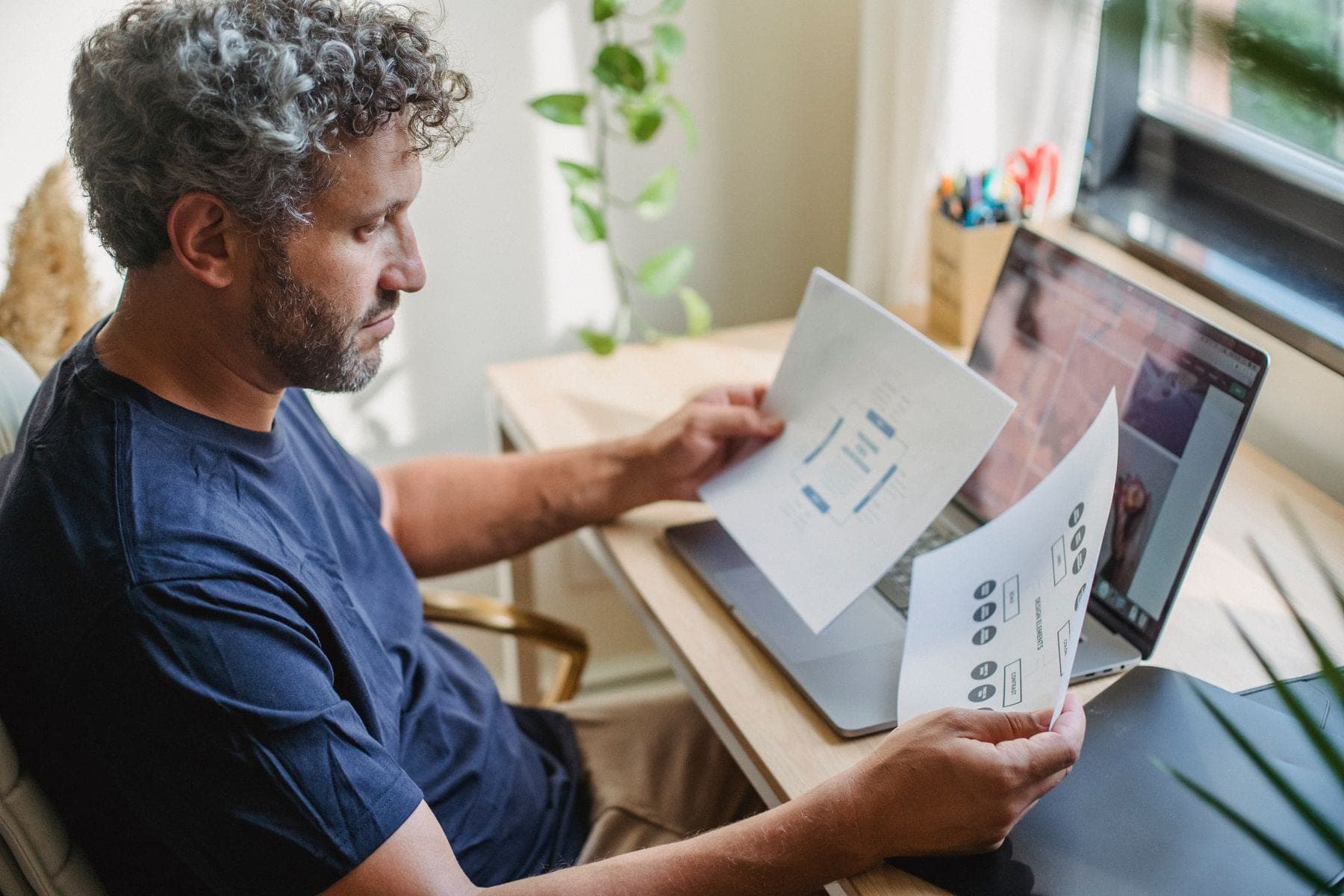 Man met baard zit aan tafel met een open laptop en leest papieren