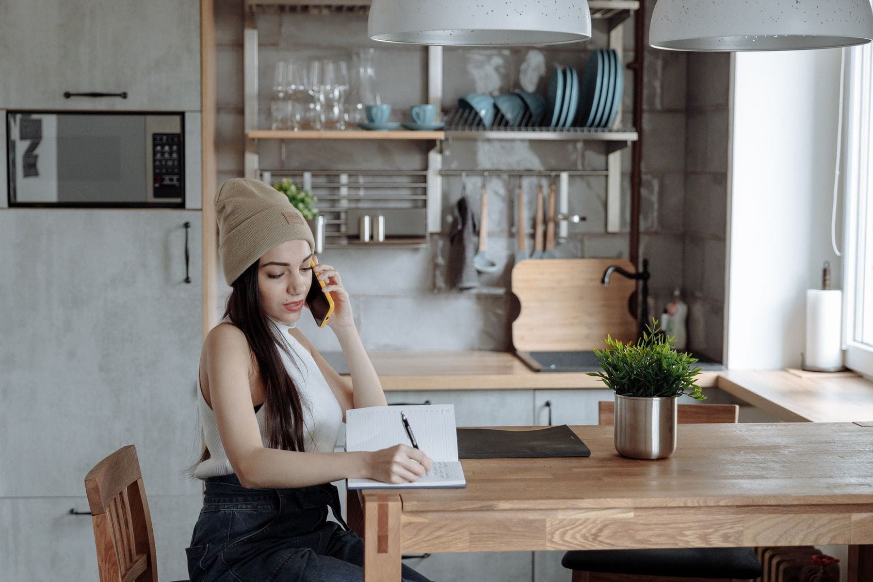Jonge vrouw met muts zit aan de keukentafel bij een opengeslagen schrift en belt met haar telefoon