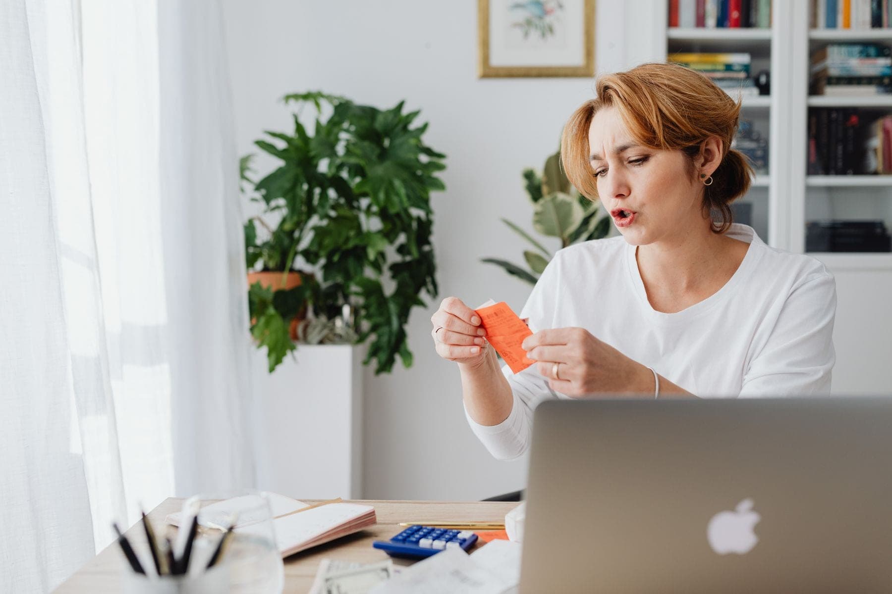 Een vrouw bekijkt rekeningen aan een bureau thuis, met een laptop en planten