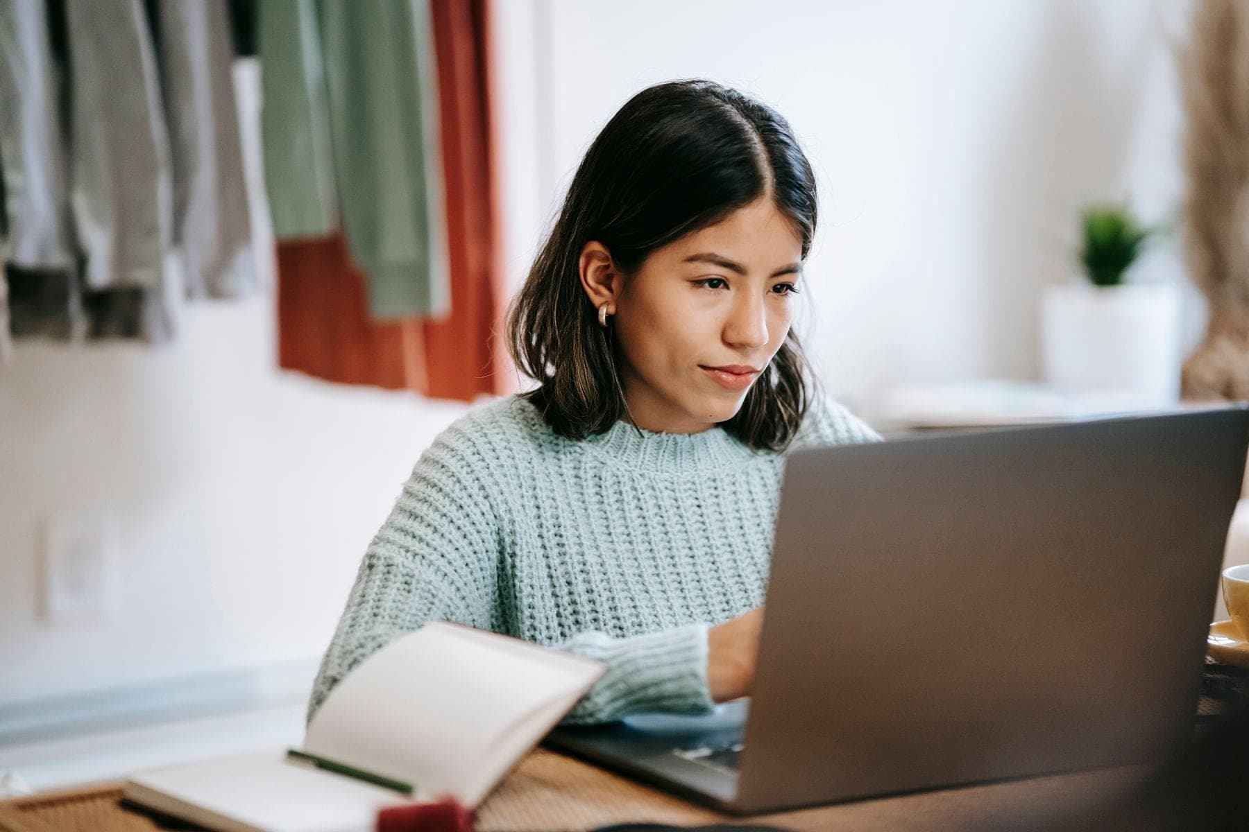 Een jonge vrouw studeert thuis aan tafel op een laptop, met een notitieblok en pen ernaast