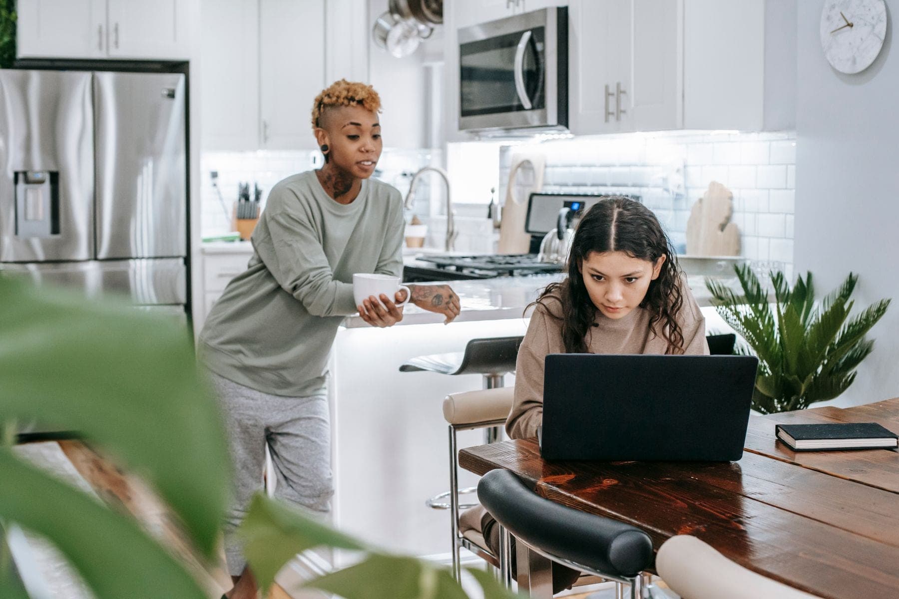 Twee jonge vrouwen aan de keukentafel, een werkt op een laptop terwijl de ander koffie drinkt
